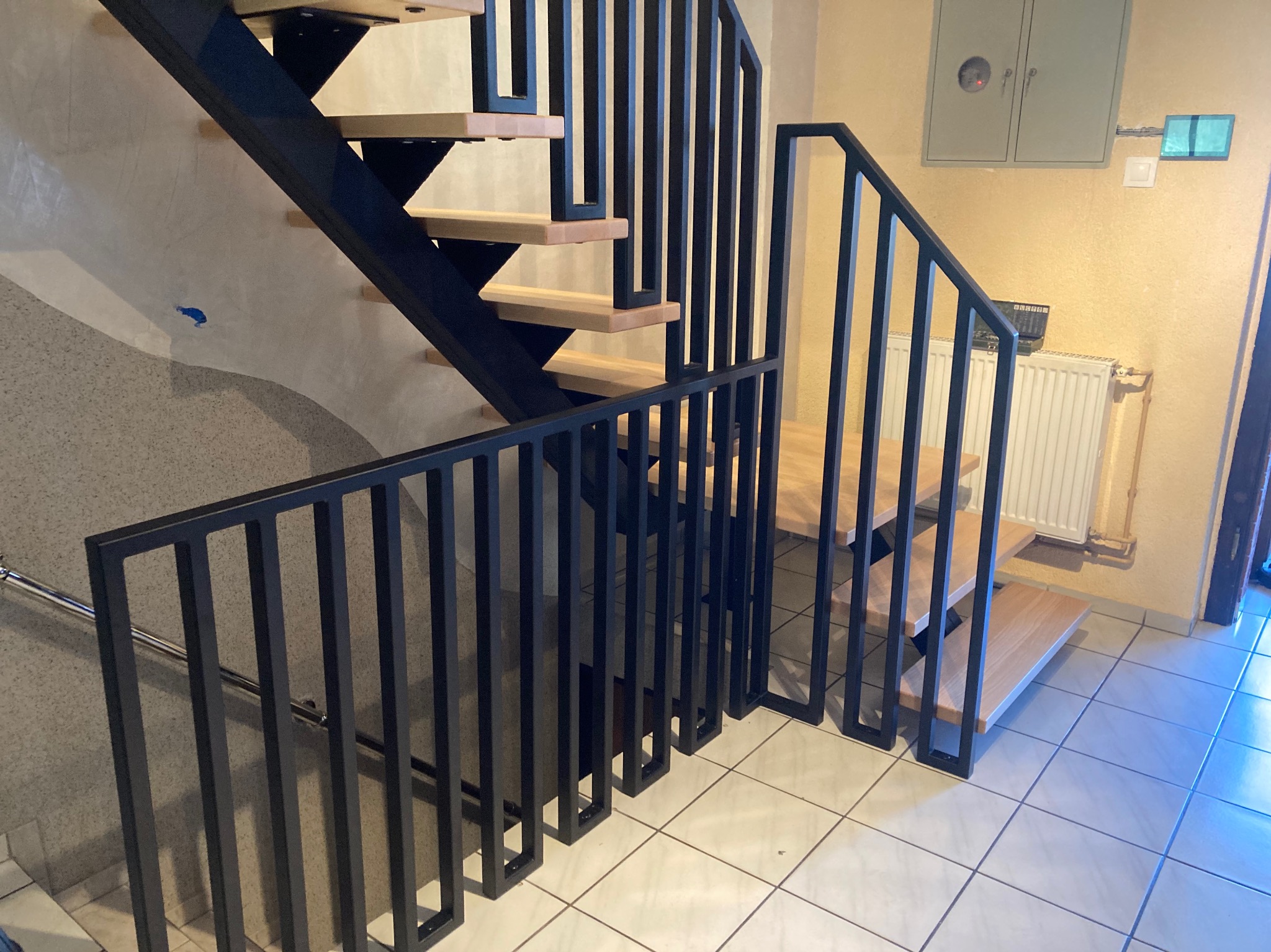 Nowoczesne schody z czarną metalową konstrukcją i drewnianymi stopniami w jasnym wnętrzu, widoczna balustrada z pionowych prętów.