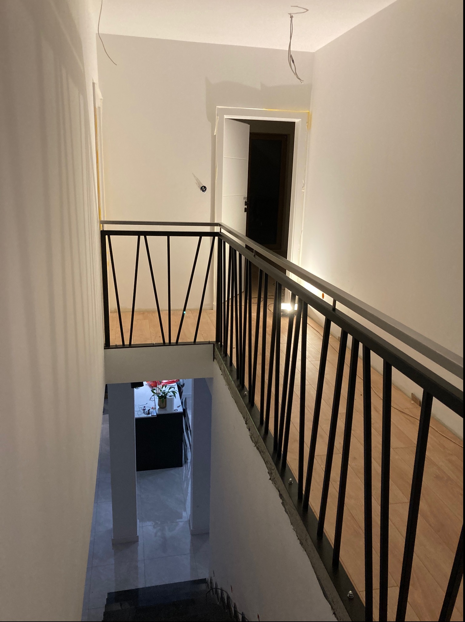 Wnętrze w trakcie wykańczania, widok z góry na schody z czarną metalową balustradą o pionowych szczeblach, drewnopodobna podłoga na górnym poziomie, w tle widoczne pomieszczenie na dole z białymi...