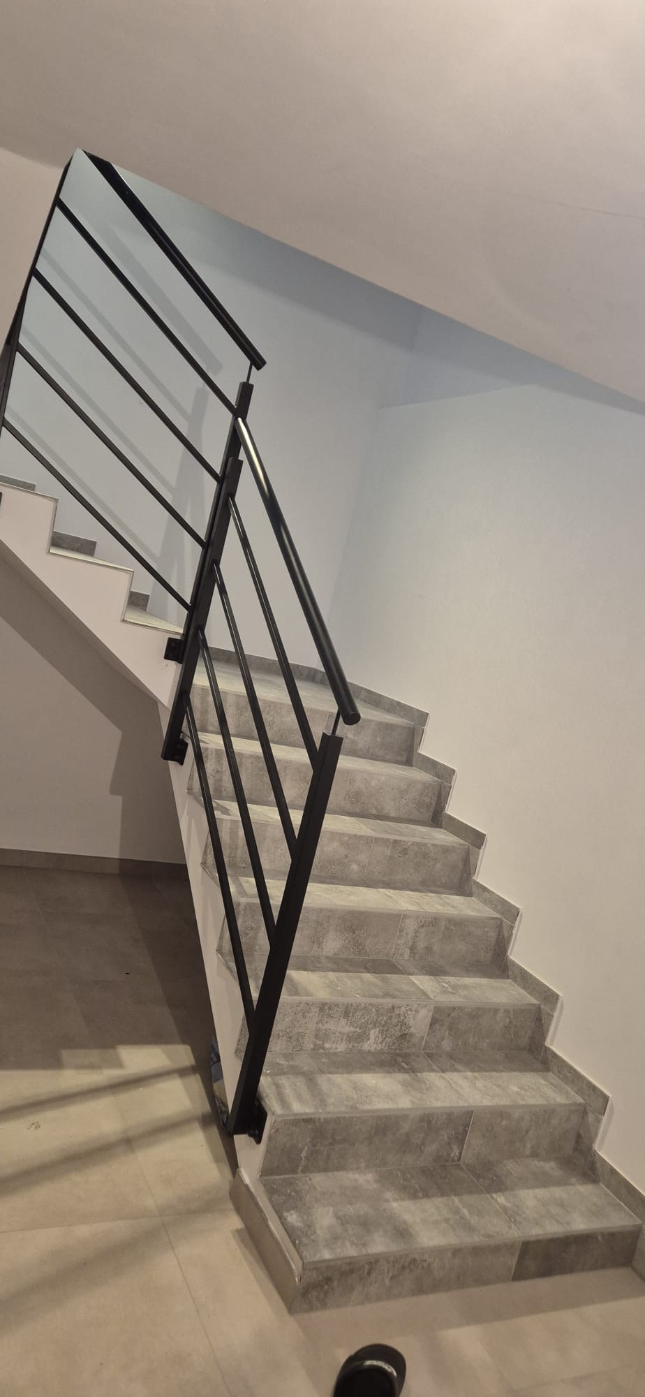 Schody wewnętrzne z szarymi płytkami i czarną, metalową balustradą o minimalistycznym designie. Widok z dołu, skupienie na detalach wykończenia.