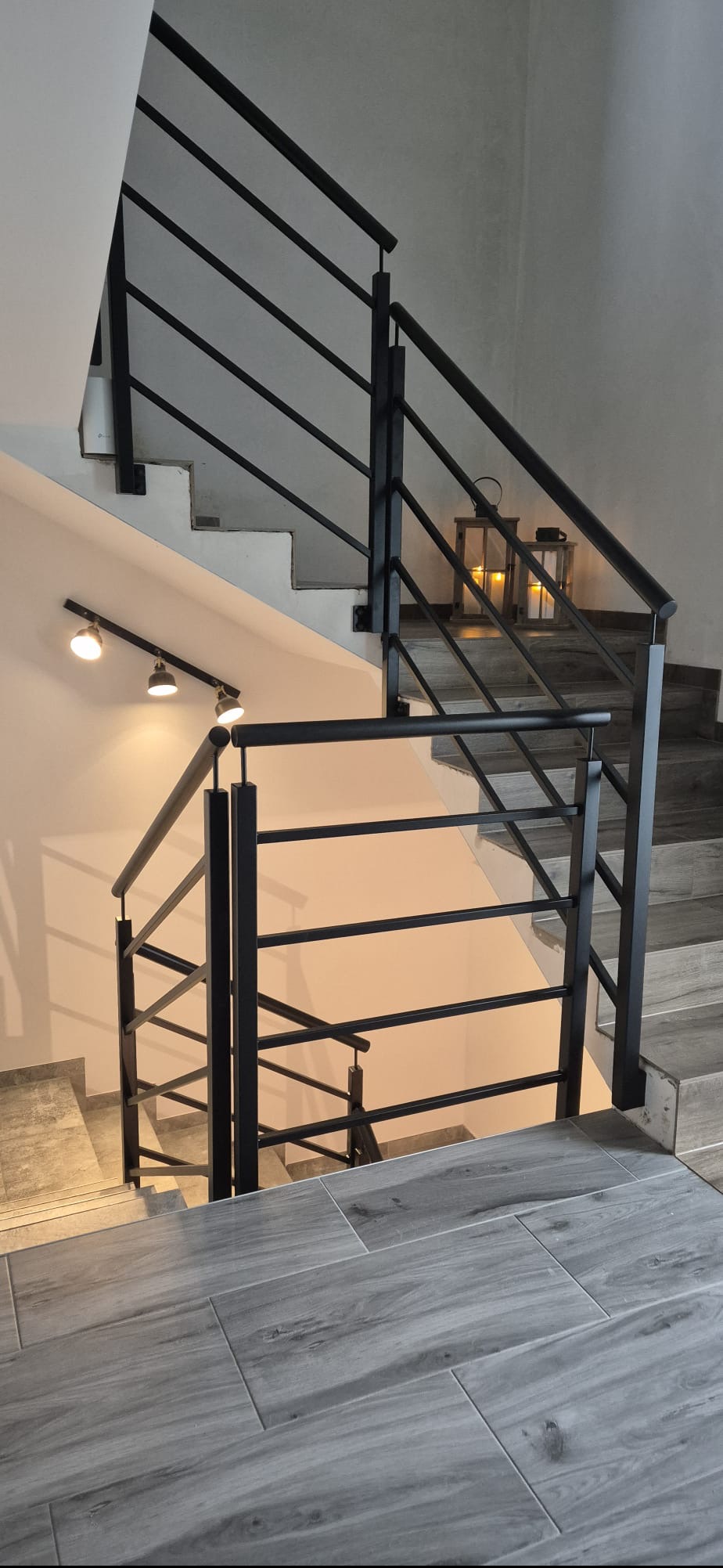 Nowoczesne schody z czarną, metalową balustradą i drewnopodobnymi płytkami. Dwa lampiony na schodach dodają ciepłego światła. Oświetlenie punktowe na ścianie.