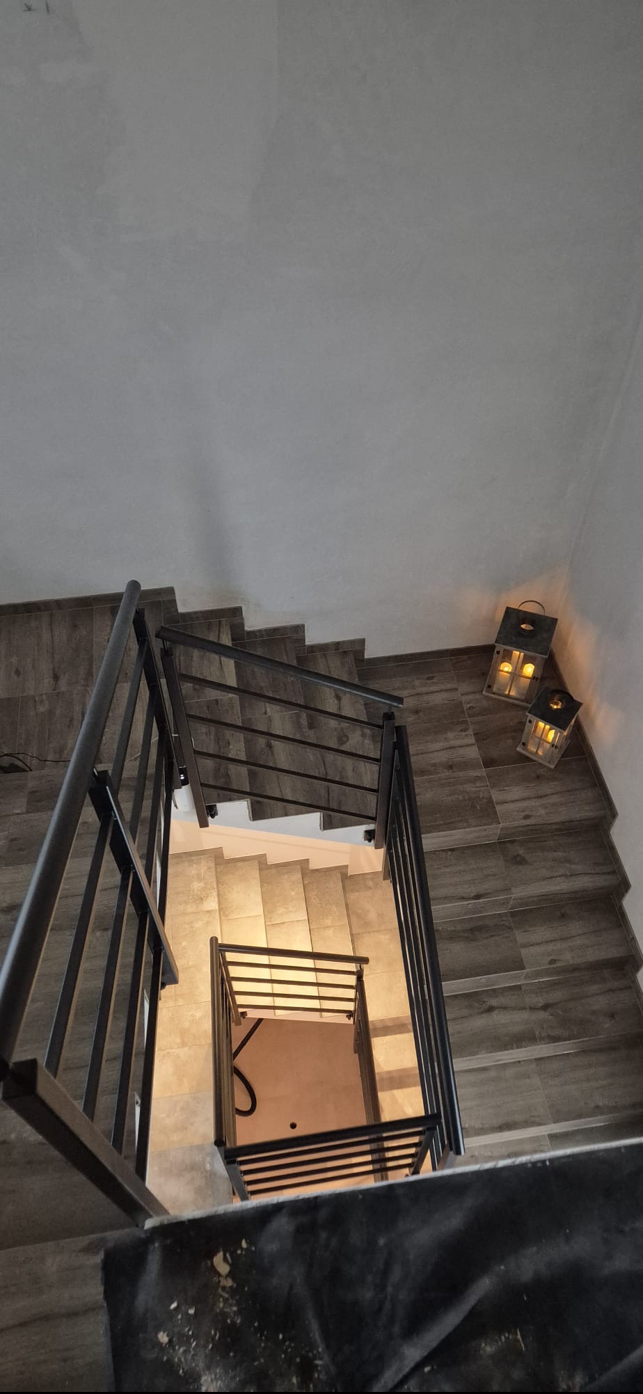 Metalowe schody z czarną balustradą, widok z góry, oświetlone lampionami. Ciemne płytki imitujące drewno na schodach.