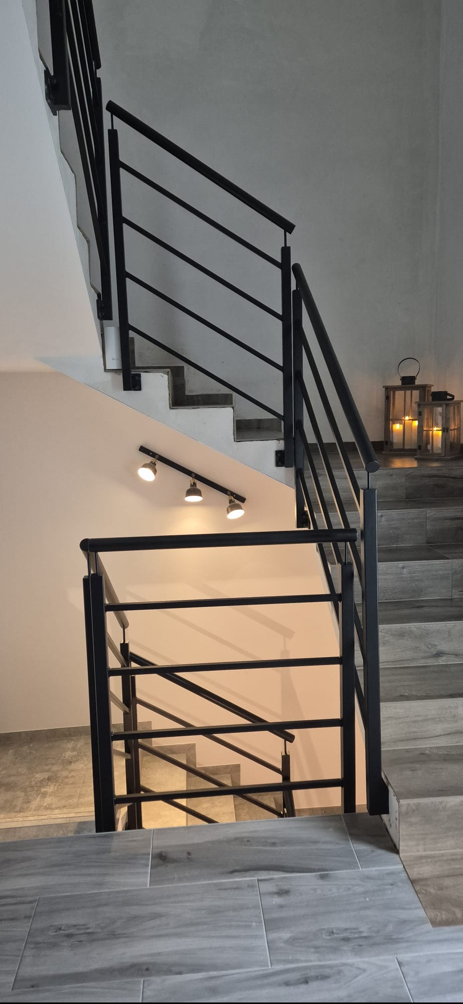 Wnętrze z metalowymi schodami i czarną, nowoczesną balustradą. Na schodach ustawione lampiony, oświetlenie punktowe na ścianie. Widok z dołu na górę.