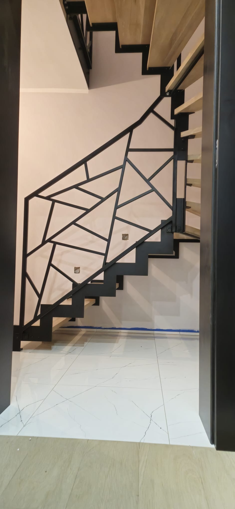 Nowoczesne schody z drewnianymi stopniami i geometryczną, czarną balustradą, widziane z dołu przez otwarte drzwi. Biała podłoga i jasna ściana w tle.