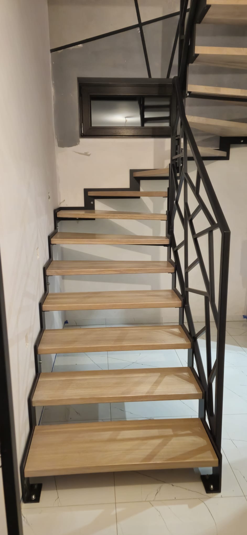 Schody metalowe z drewnianymi stopniami i designerską balustradą z czarnego metalu, widok z dołu, w tle okno i fragment ściany.