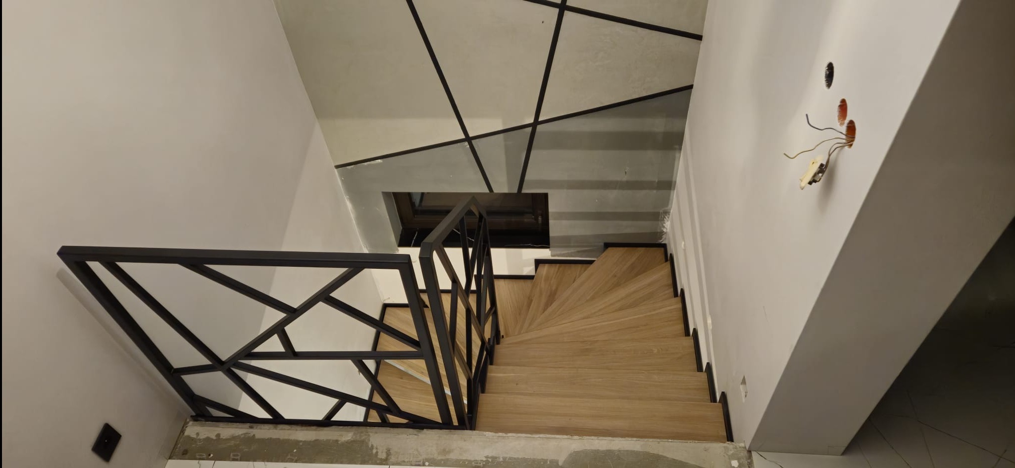 Nowoczesne schody drewniane z czarną, geometryczną balustradą metalową, widziane z góry. Na suficie widoczny wzór z czarnych linii. Otwory na gniazdka elektryczne na ścianie.