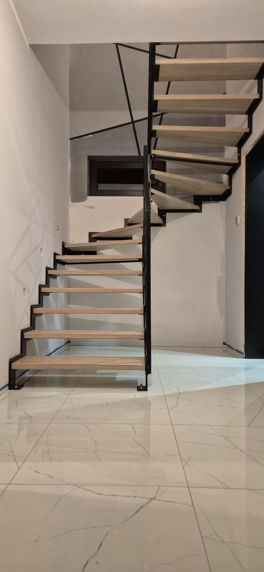 Nowoczesne schody z drewnianymi stopniami i czarną metalową konstrukcją na białych marmurowych płytkach. W tle surowa ściana z oknem i geometrycznym wzorem.