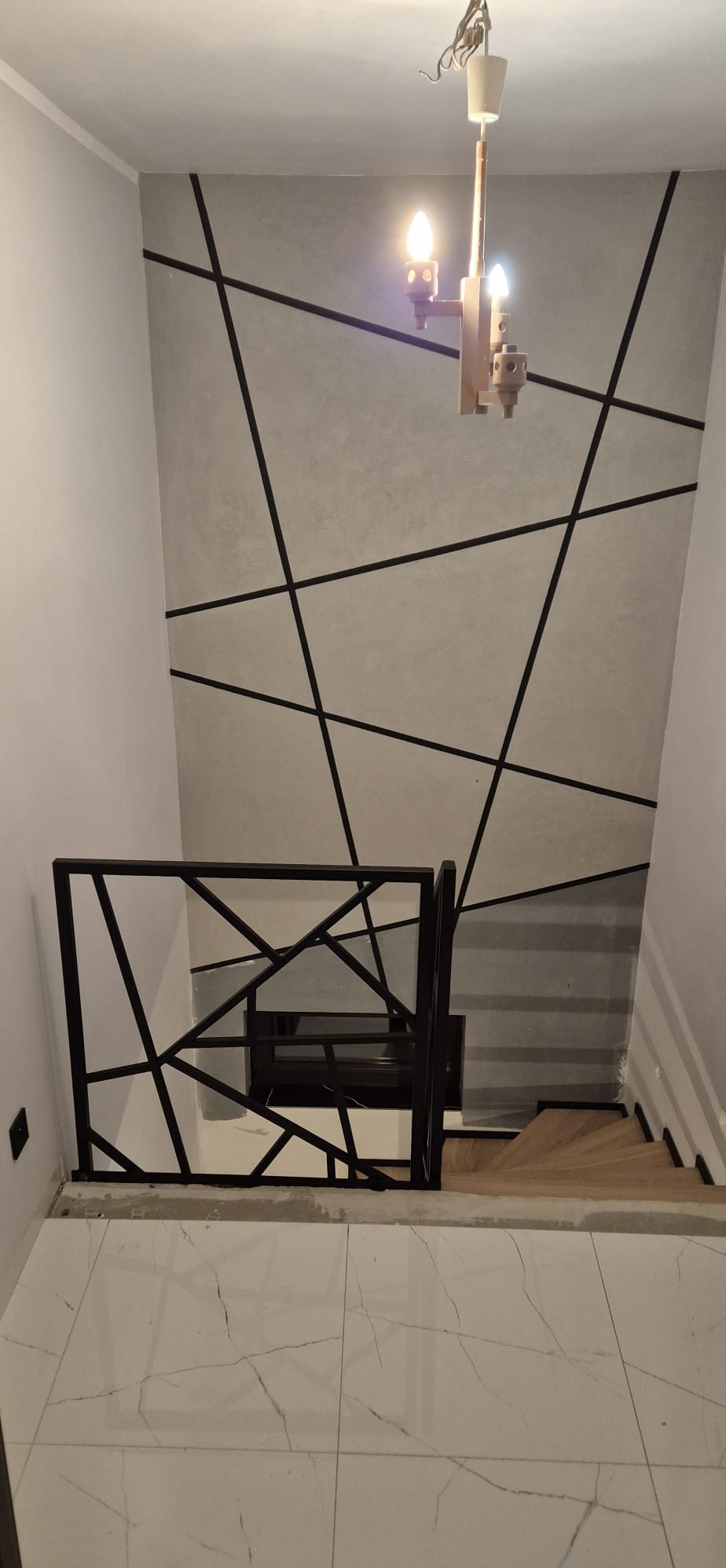 Nowoczesna balustrada schodowa z czarnych profili o geometrycznym wzorze, widok z góry na białą marmurową podłogę i drewniane schody, ściana z dekoracyjnymi liniami.
