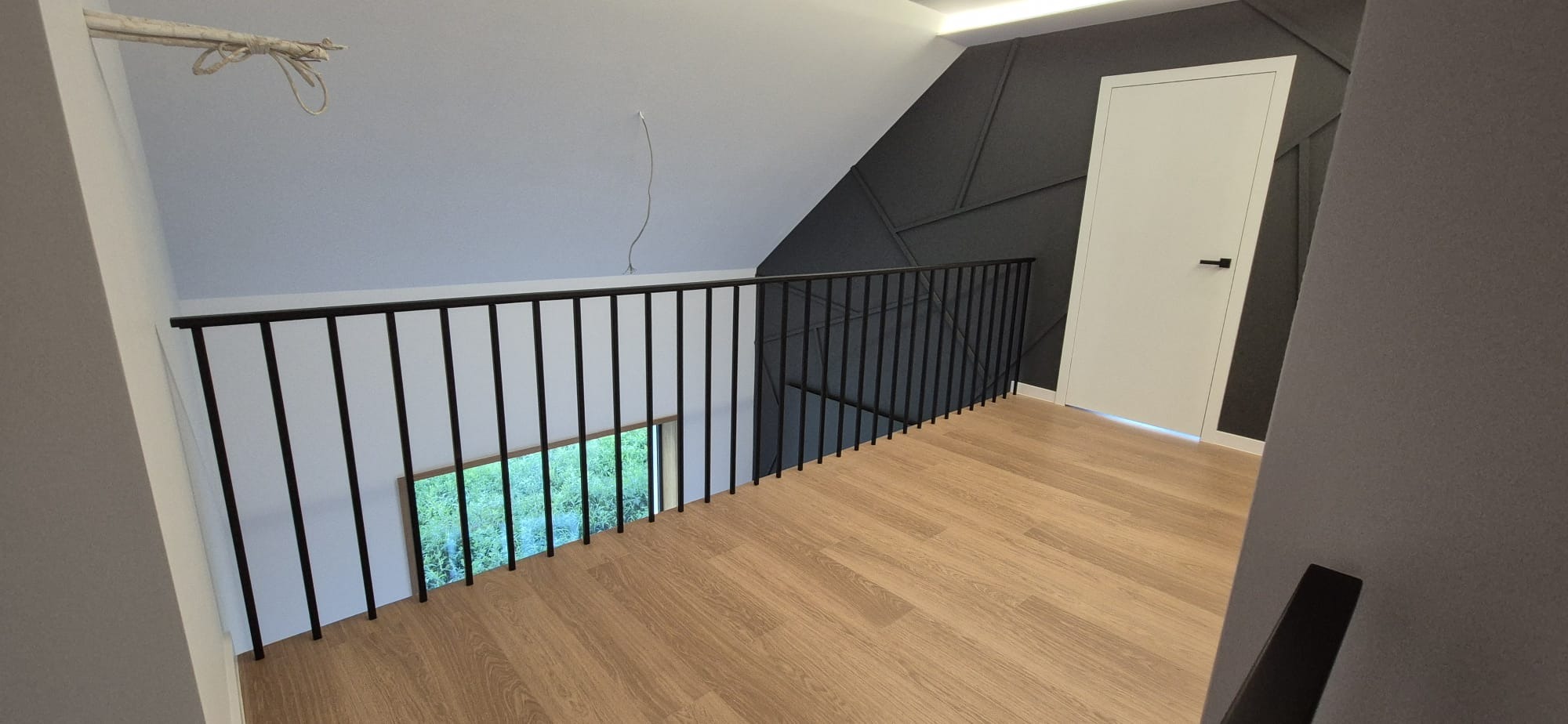 Wnętrze poddasza z drewnianą podłogą i czarną, metalową balustradą. Widoczne okno z widokiem na zieleń i dekoracyjna, ciemna ściana z geometrycznym wzorem.