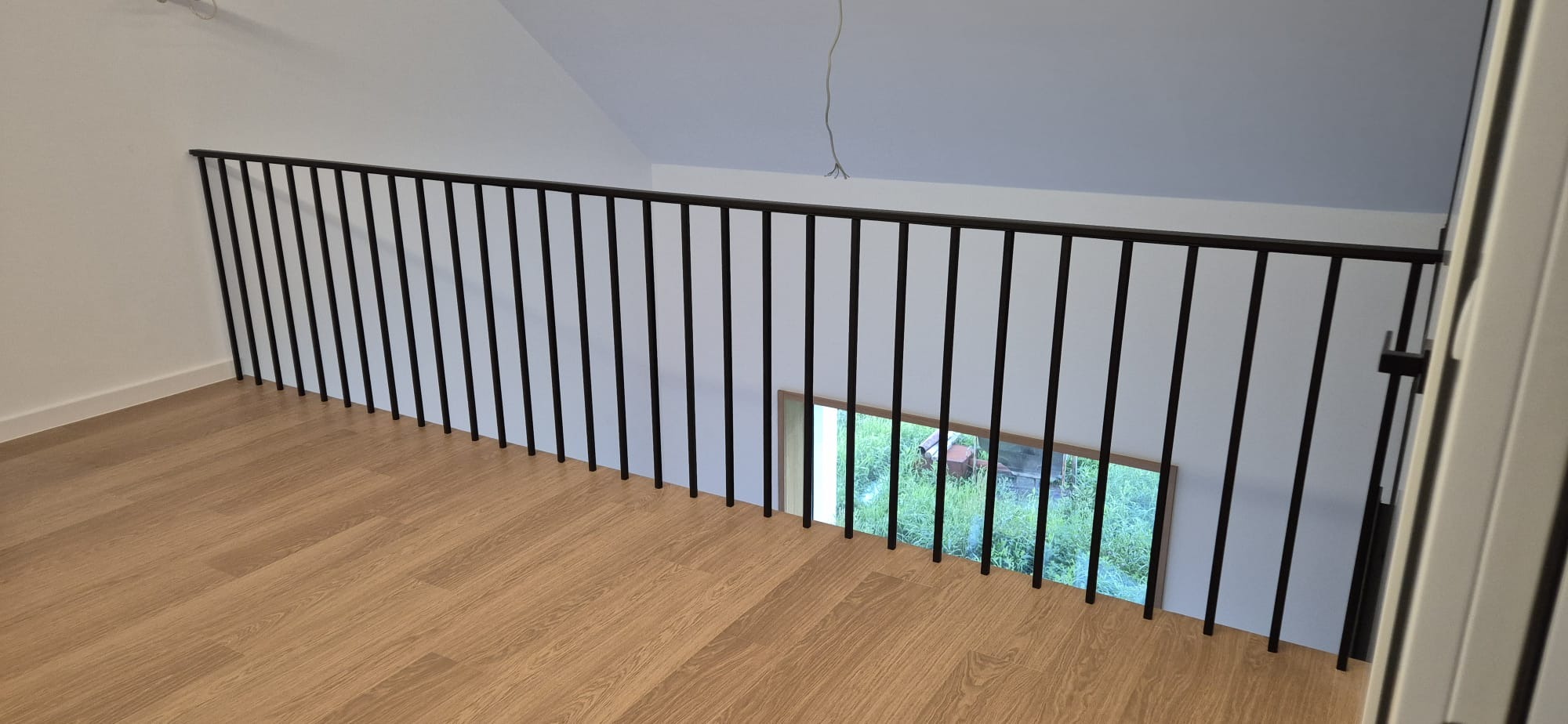 Wnętrze z drewnianą podłogą i czarną, minimalistyczną balustradą. Widok na niższy poziom przez otwór w podłodze. Nowoczesny design, jasne ściany.