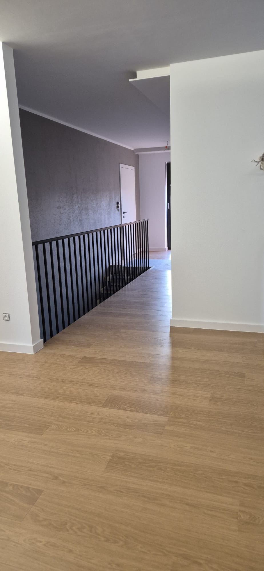 Nowoczesne wnętrze z drewnianą podłogą i czarną balustradą schodową. Szara ściana w tle dodaje elegancji. Widok z góry na korytarz.