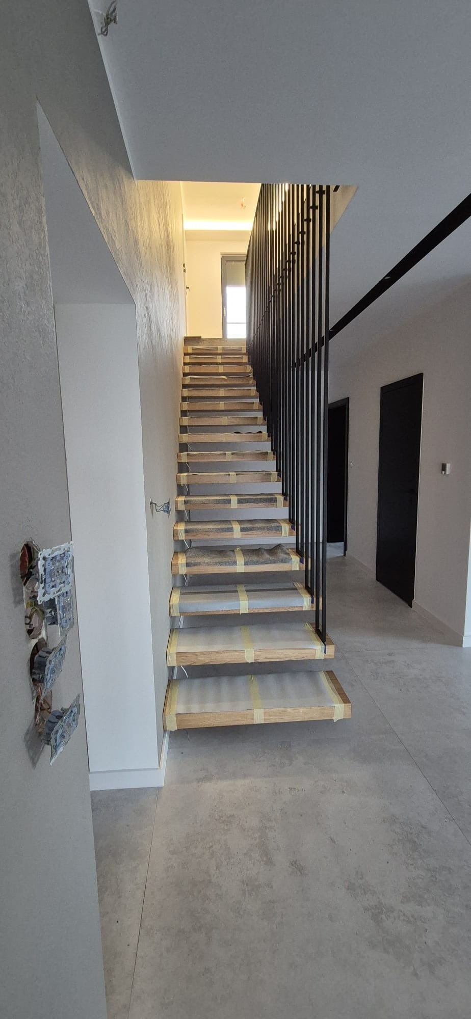Wnętrze domu z drewnianymi schodami zabezpieczonymi folią i taśmą, z czarną, pionową balustradą. Widoczne niedokończone gniazdka elektryczne na ścianie.