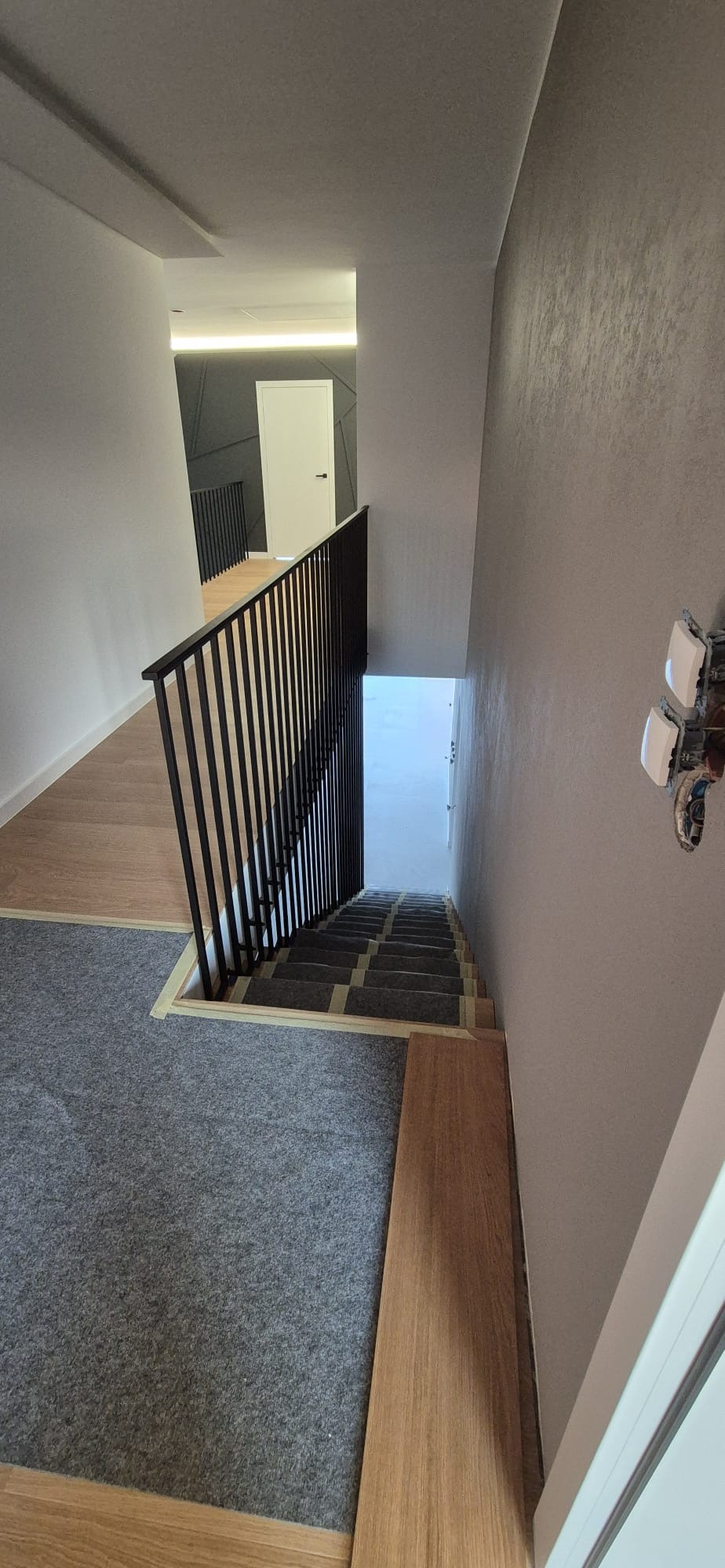 Wnętrze domu z widokiem na schody z czarną, pionową balustradą. Część schodów wyłożona szarą wykładziną, a część drewnem. Widoczne niedokończone prace wykończeniowe.