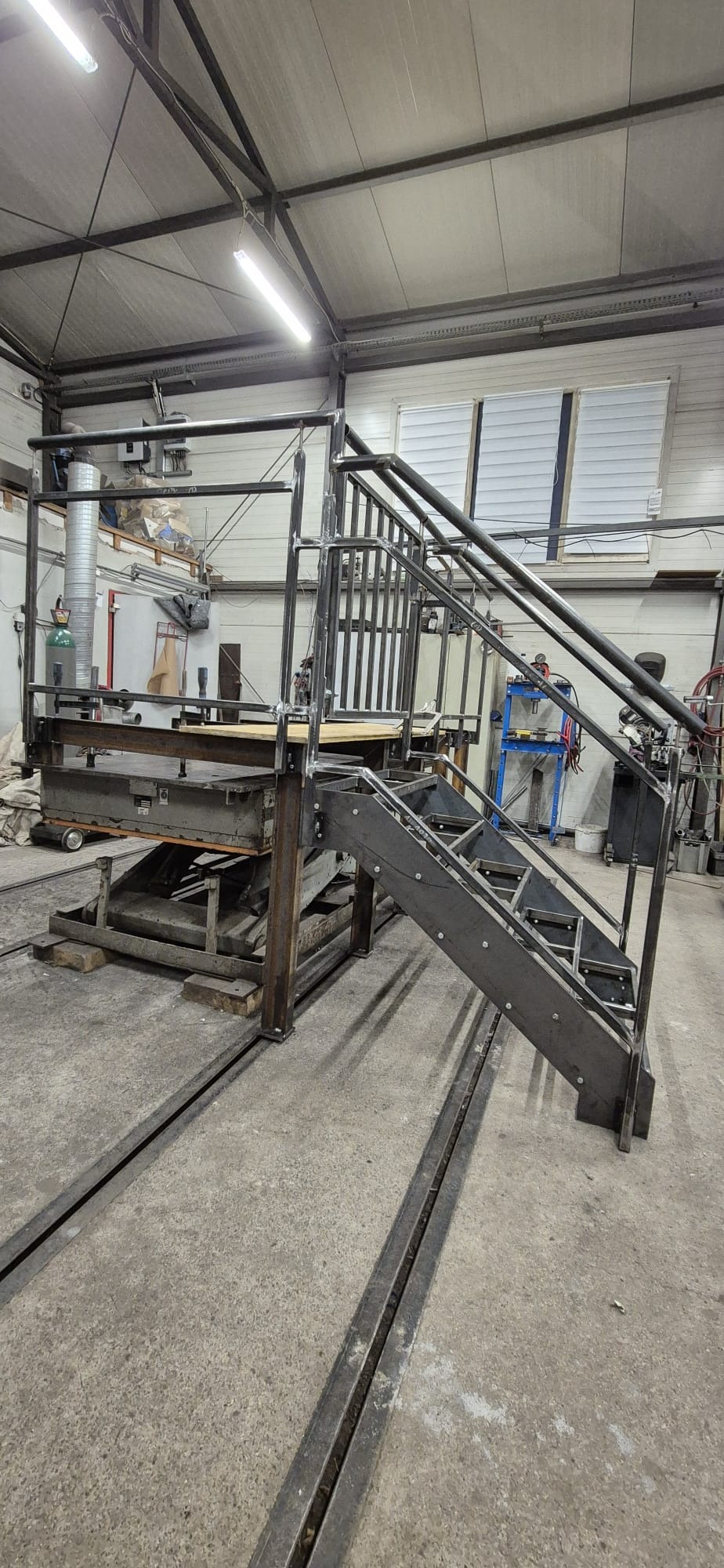 Konstrukcja metalowa z schodami i balustradą w hali produkcyjnej, widoczne ślady spawania. Metalowa platforma na podnośniku, oświetlenie jarzeniowe w tle.