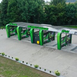 Mariusz Drążewski EKOWASH - Widok z góry na nowoczesną, bezdotykową myjnię samochodową z zielonymi elementami konstrukcyjnymi i napisem 'BEZDOTYKOWA 24H', otoczoną zielenią i kostką brukową.