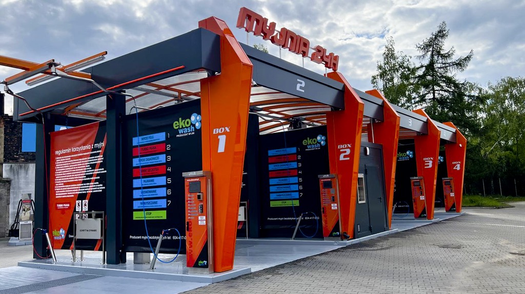 Zewnętrzny widok na samoobsługową myjnię samochodową z czterema stanowiskami, wykończoną w kolorach czarnym i pomarańczowym, z widocznym cennikiem usług na każdym stanowisku.