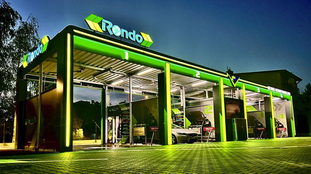 Wieczorny widok na nowoczesną, oświetloną myjnię samochodową Rondo z czterema stanowiskami, z których jedno zajęte jest przez srebrny samochód osobowy.