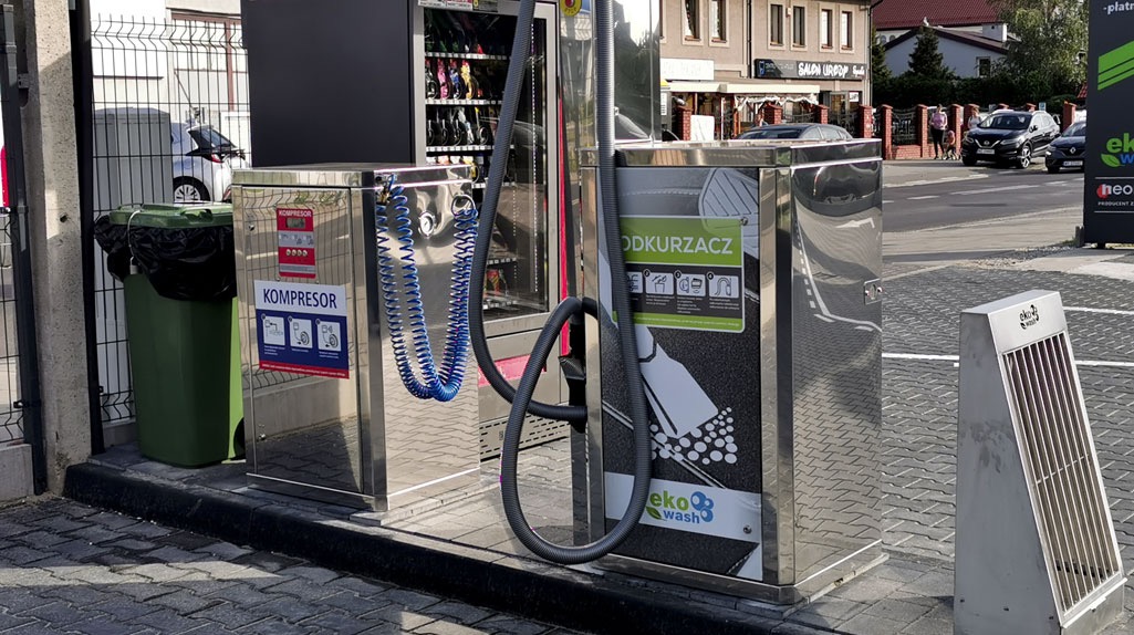 Samoobsługowa stacja mycia pojazdów z kompresorem i odkurzaczem, widoczny kosz na śmieci i automat z akcesoriami do mycia, na ulicy w tle budynki.