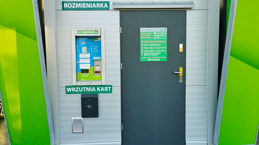 Wejście do samoobsługowej myjni samochodowej 24h z automatem do rozmieniania pieniędzy i wrzutnią na karty płatnicze, obok drzwi informacja o możliwości płatności kartą rabatową.