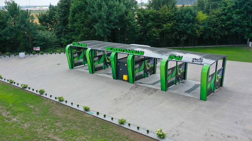 Widok z góry na nowoczesną, bezdotykową myjnię samochodową z zielonymi elementami konstrukcyjnymi i napisem 'BEZDOTYKOWA 24H', otoczoną zielenią i kostką brukową.