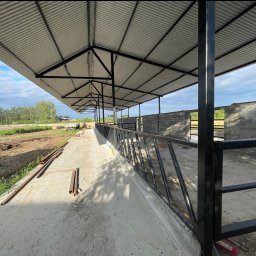 Dachy Twardysko - Konstrukcja z blachy trapezowej wsparta na stalowych słupach, w tle budynek z bloczków betonowych i krajobraz wiejski.