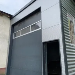 Nowoczesny garaż blaszany z segmentową bramą garażową w kolorze grafitowym, widoczne elementy konstrukcyjne z blachy trapezowej, częściowo otwarta brama wejściowa z boku.