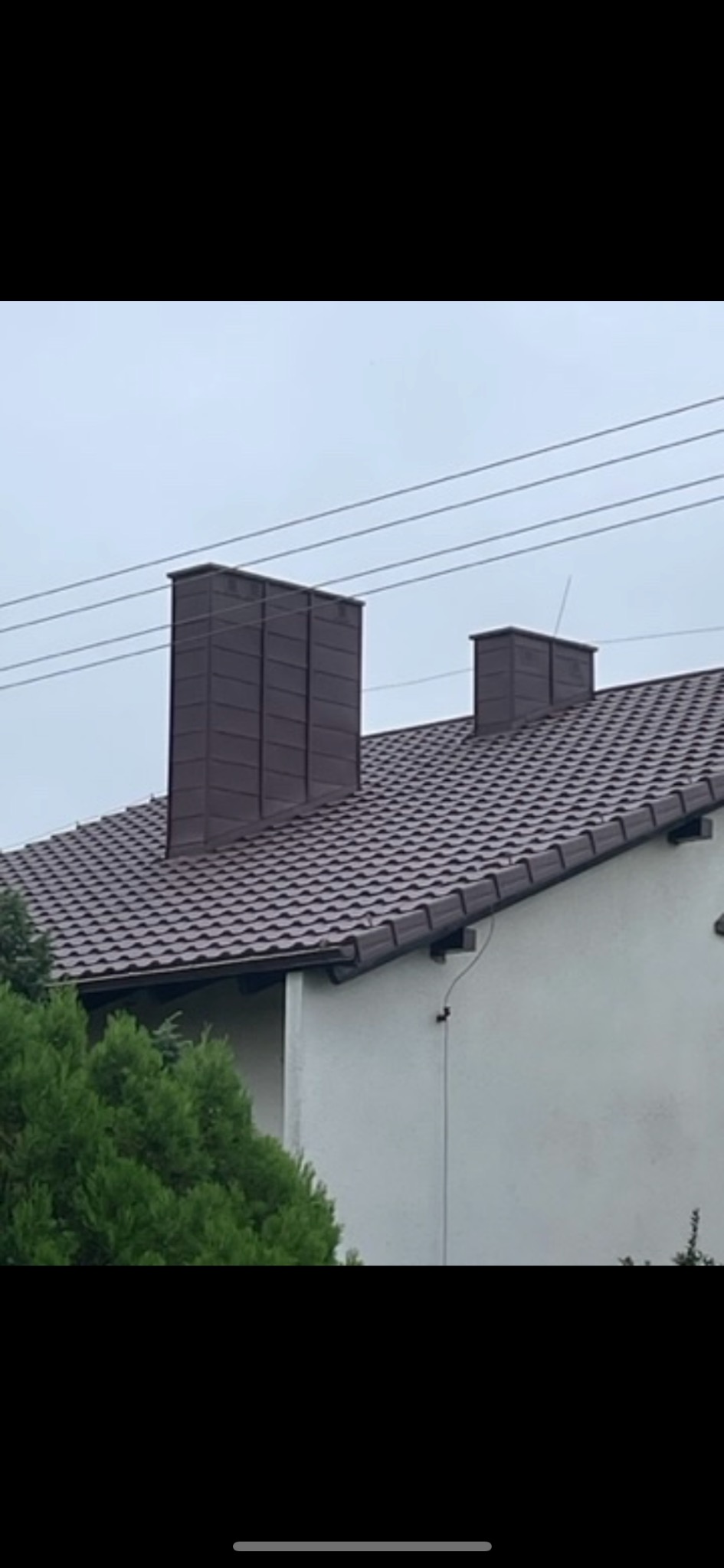 Brązowa dachówka ceramiczna na dachu domu jednorodzinnego z dwoma kominami, jeden wysoki, drugi niższy, na tle pochmurnego nieba.