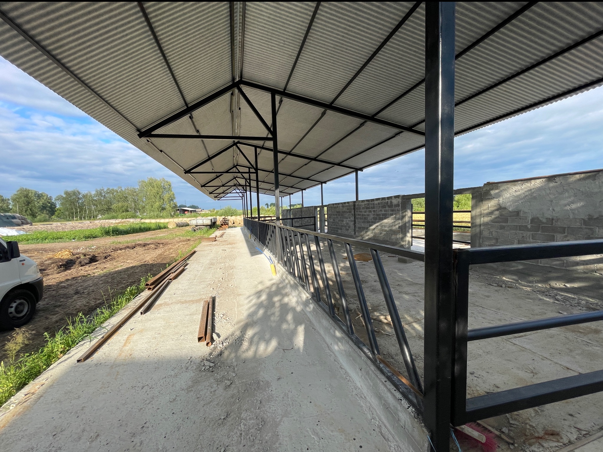 Konstrukcja z blachy trapezowej wsparta na stalowych słupach, w tle budynek z bloczków betonowych i krajobraz wiejski.
