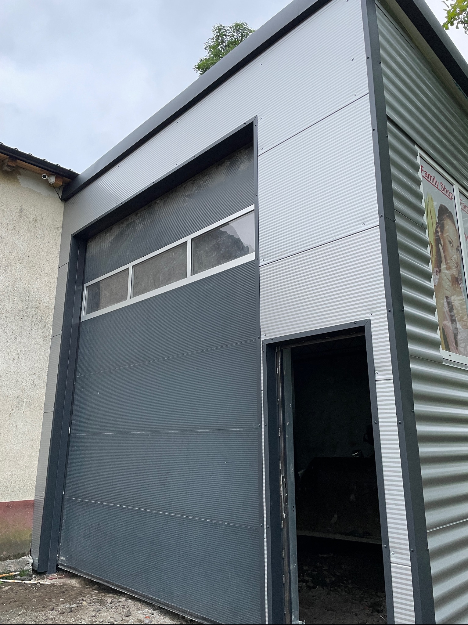 Nowoczesny garaż blaszany z segmentową bramą garażową w kolorze grafitowym, widoczne elementy konstrukcyjne z blachy trapezowej, częściowo otwarta brama wejściowa z boku.