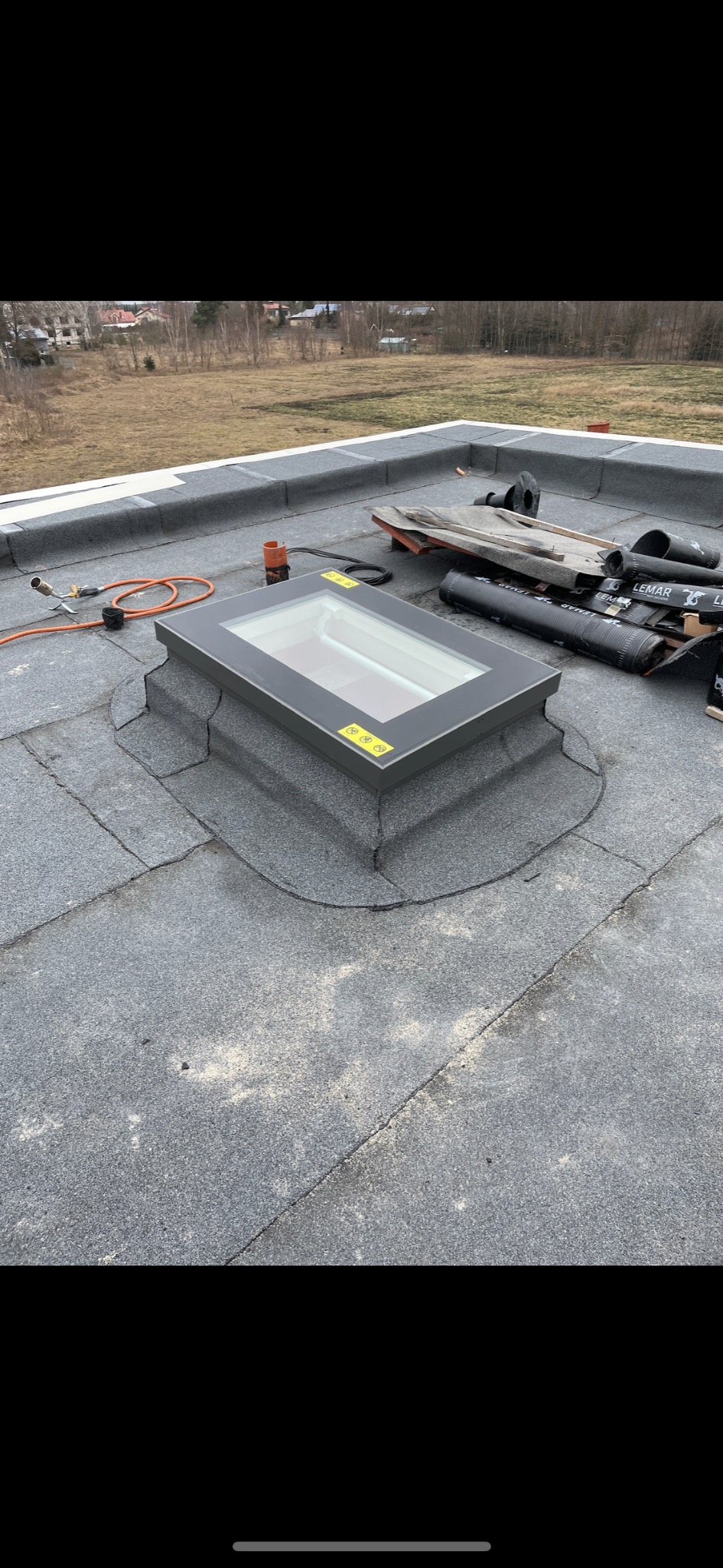 Szary dach z papy z zamontowanym oknem dachowym, widoczne narzędzia i materiały budowlane, w tle zabudowa jednorodzinna.