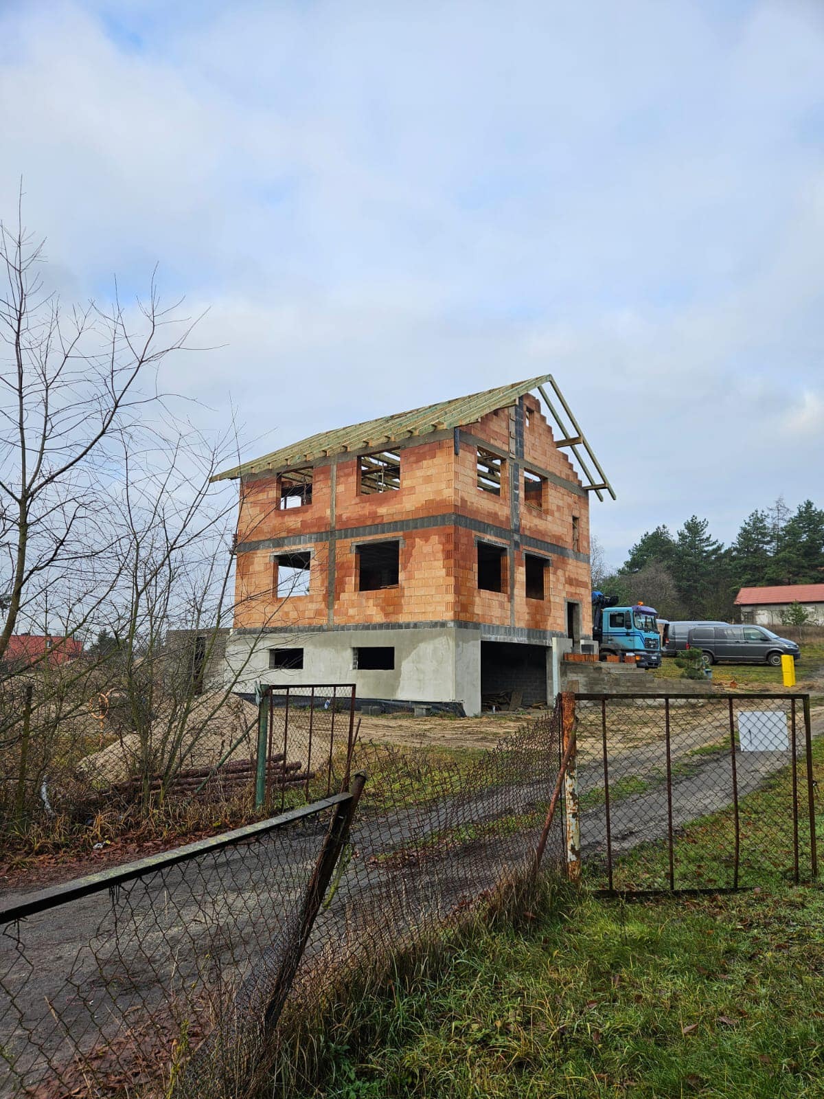 Surowy stan dwupiętrowego domu z czerwonej cegły z widoczną więźbą dachową i niezabezpieczonymi otworami okiennymi, częściowo otynkowany fundament, na tle pochmurnego nieba i drzew