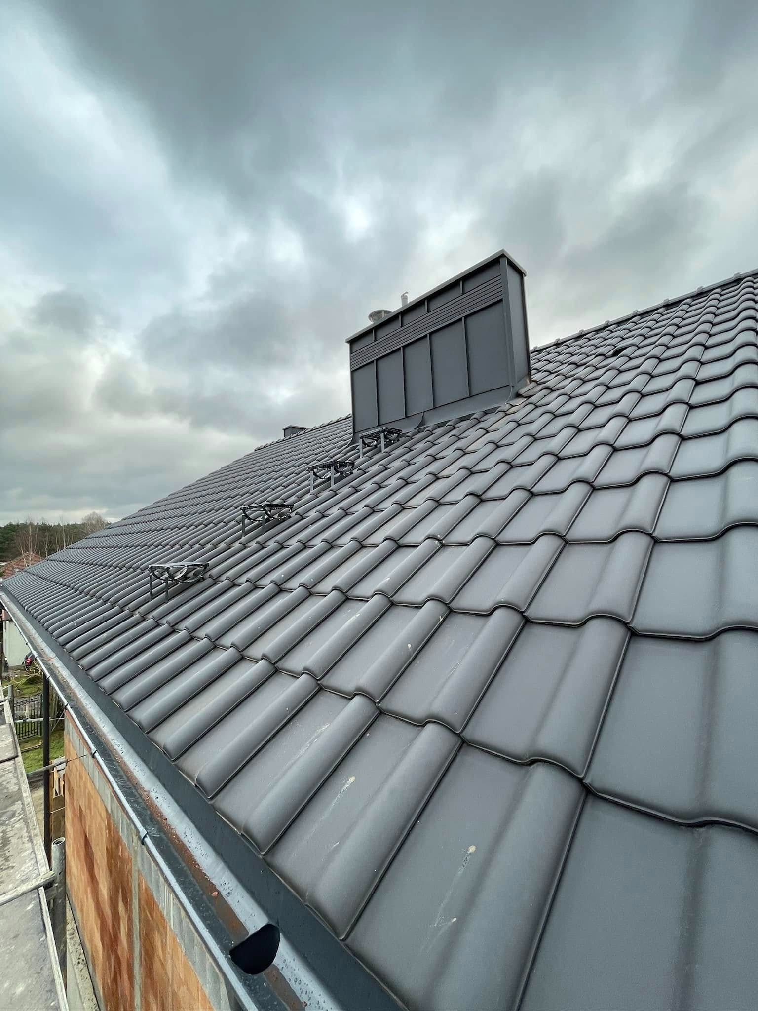 Nowy, grafitowy dach z ceramicznej dachówki karpiówki z zamontowanymi stopniami kominiarskimi i obróbką komina w kolorze dachu, widok z dołu na tle pochmurnego nieba.