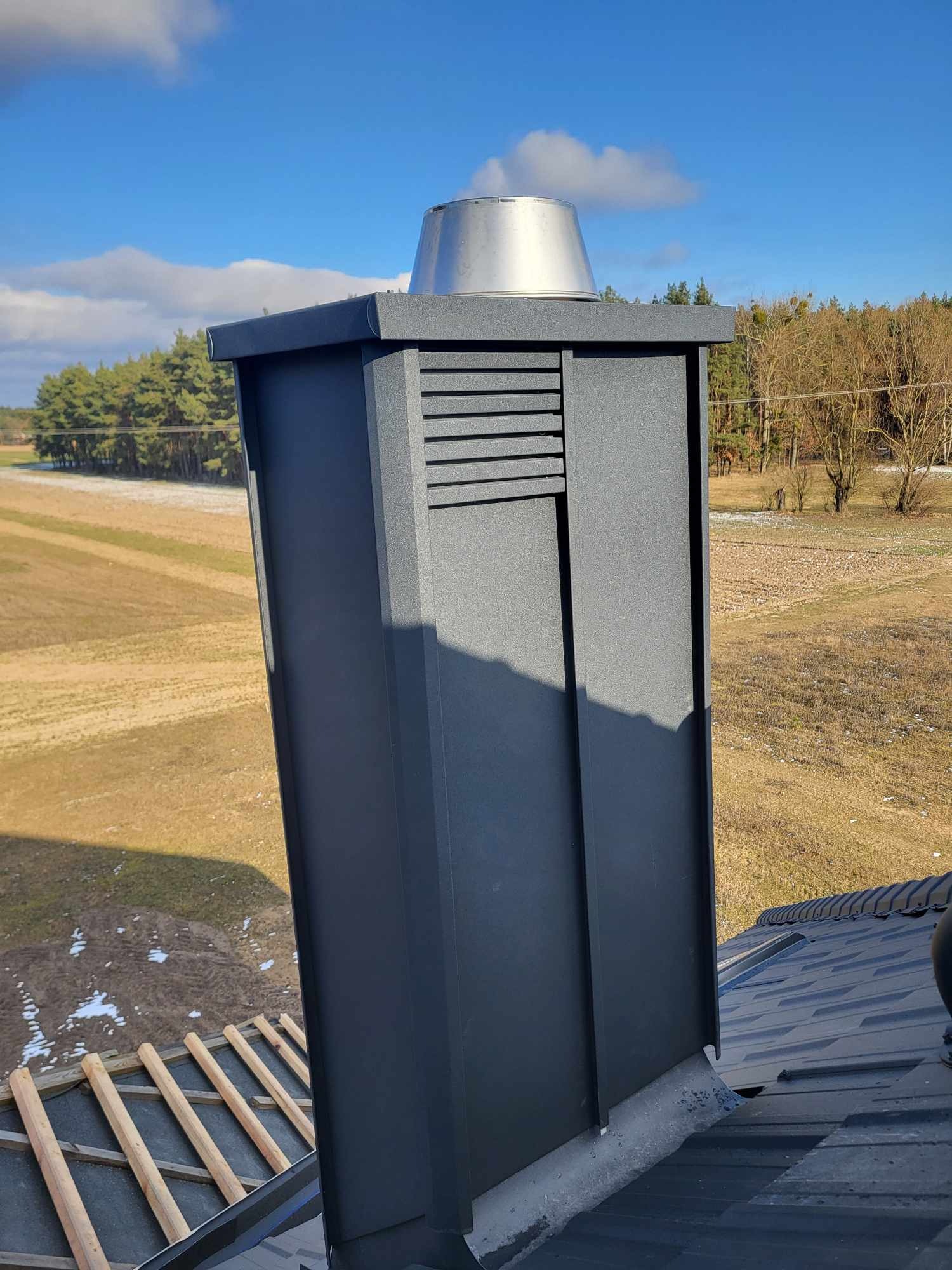 Nowoczesny, grafitowy komin wentylacyjny z metalowym daszkiem na dachu z blachy na tle zaśnieżonego pola i lasu w słoneczny dzień.