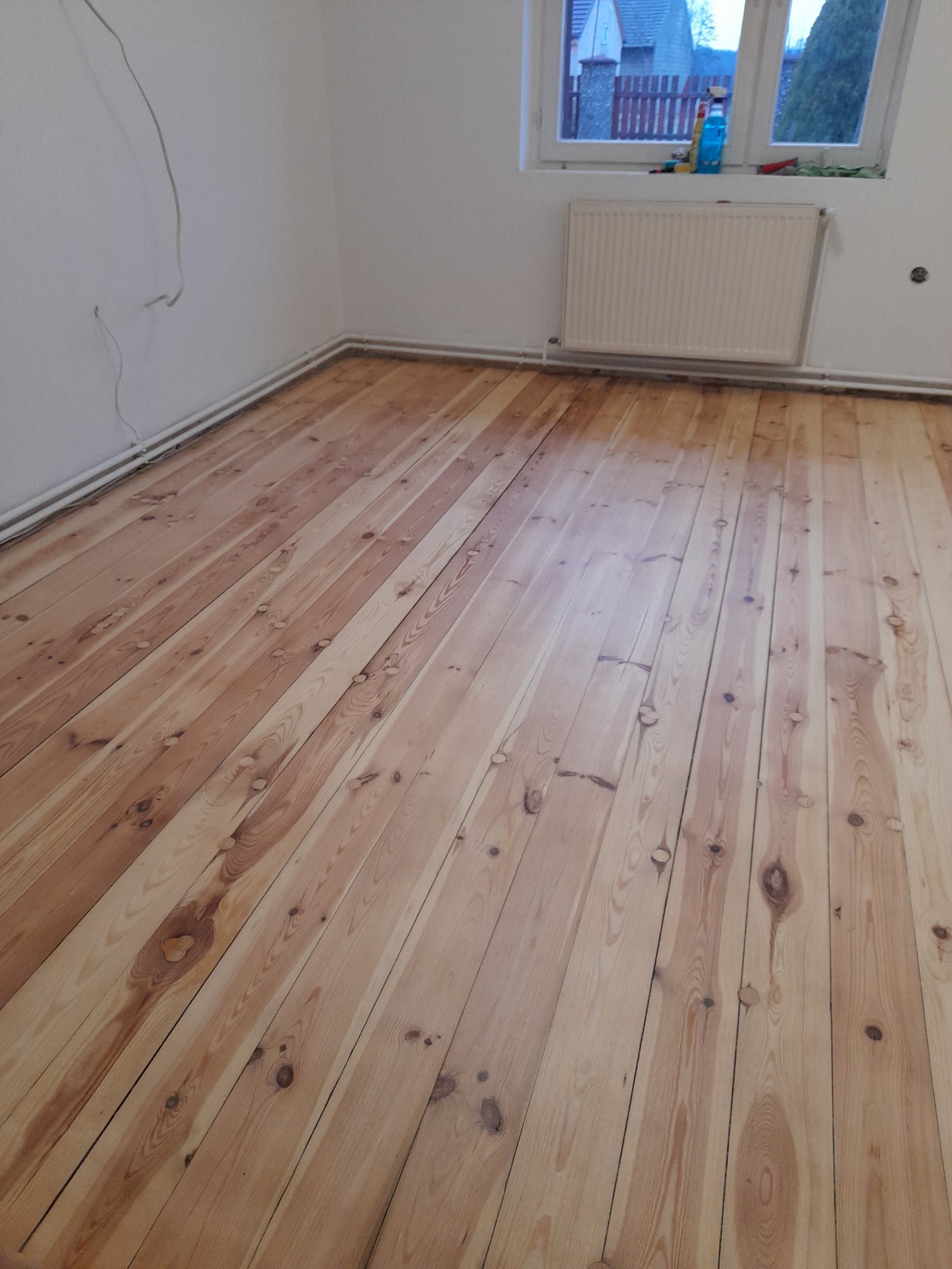 Wypolerowana podłoga z desek w jasnym odcieniu drewna, widoczne ślady po cyklinowaniu, listwy przypodłogowe, okno z widokiem na dom i niebieski płyn do mycia na parapecie.