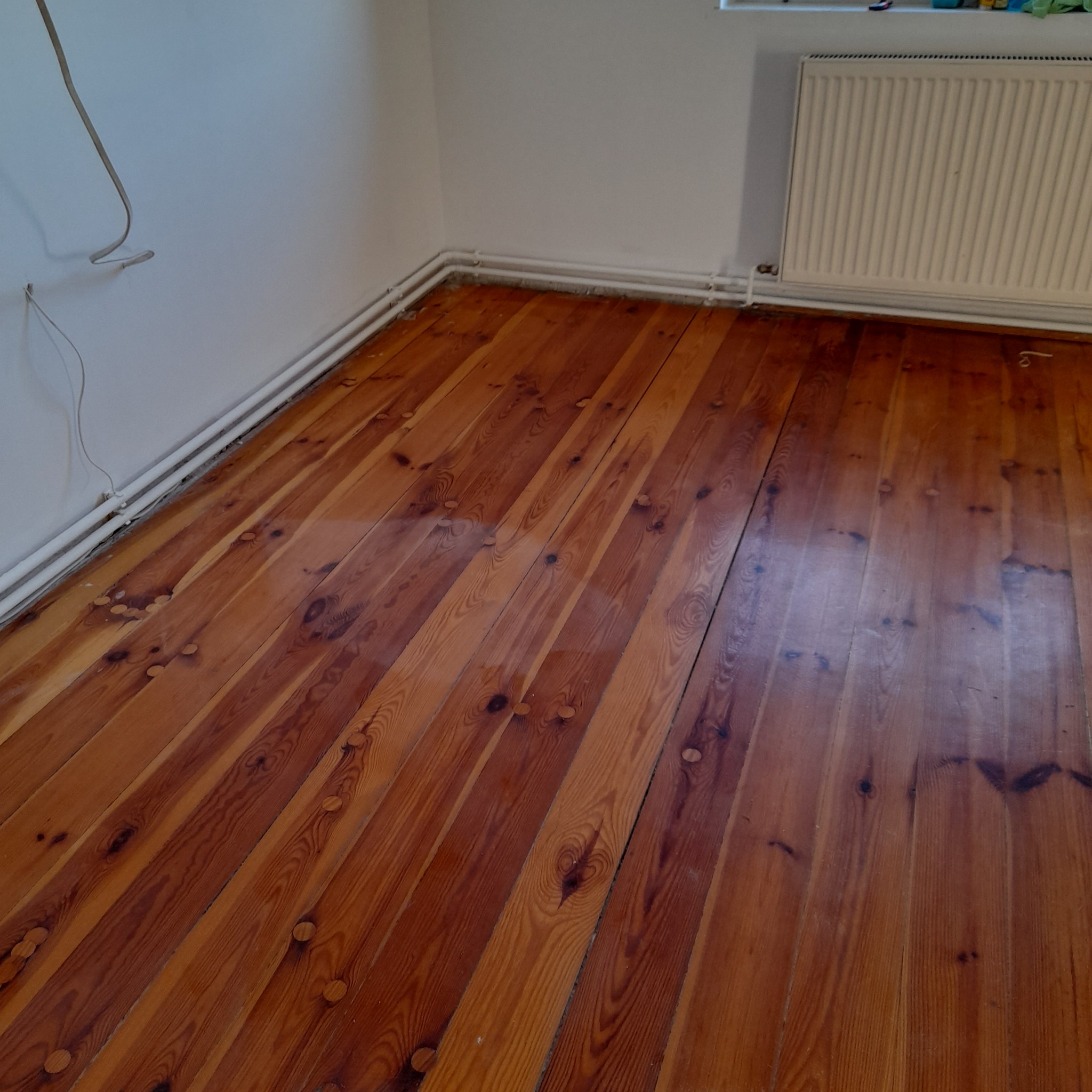 Odnowiona drewniana podłoga z widocznymi śladami po cyklinowaniu, ciepłe odcienie brązu, białe rury grzewcze przy ścianie.