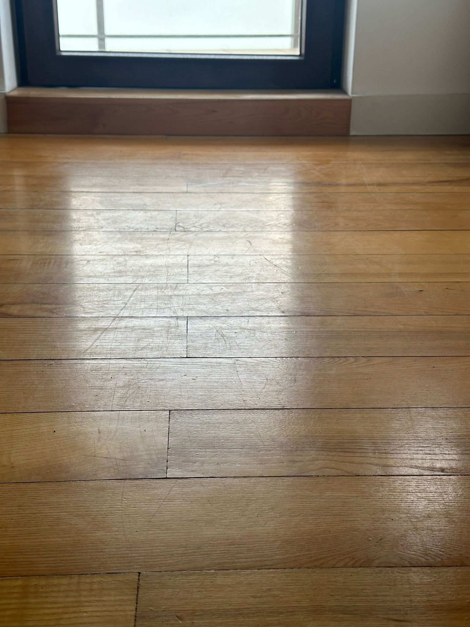 Parkiet drewniany przed oknem, widoczne rysy i ślady użytkowania, odbijające się światło podkreśla niedoskonałości.