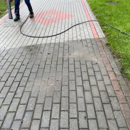 Jarosław Łukasiewicz - Częściowo umyta kostka brukowa z czerwoną obwódką, widoczny pracownik z lancą ciśnieniową i wężem. Połowa powierzchni jest mokra i czysta, druga sucha i brudna.