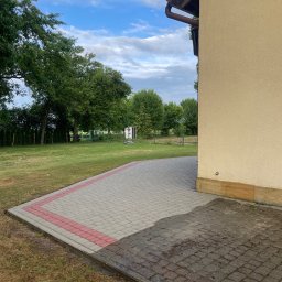 Jarosław Łukasiewicz - Jasnoszara kostka brukowa z czerwoną obwódką, częściowo mokra, przylegająca do beżowej elewacji budynku. W tle trawnik i drzewa pod błękitnym niebem.