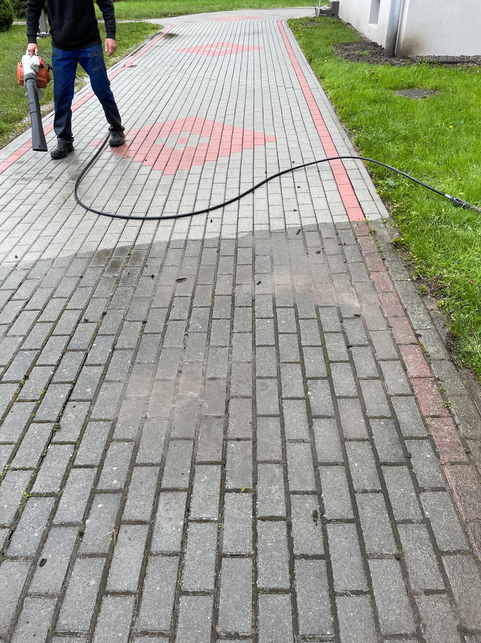 Częściowo umyta kostka brukowa z czerwoną obwódką, widoczny pracownik z lancą ciśnieniową i wężem. Połowa powierzchni jest mokra i czysta, druga sucha i brudna.