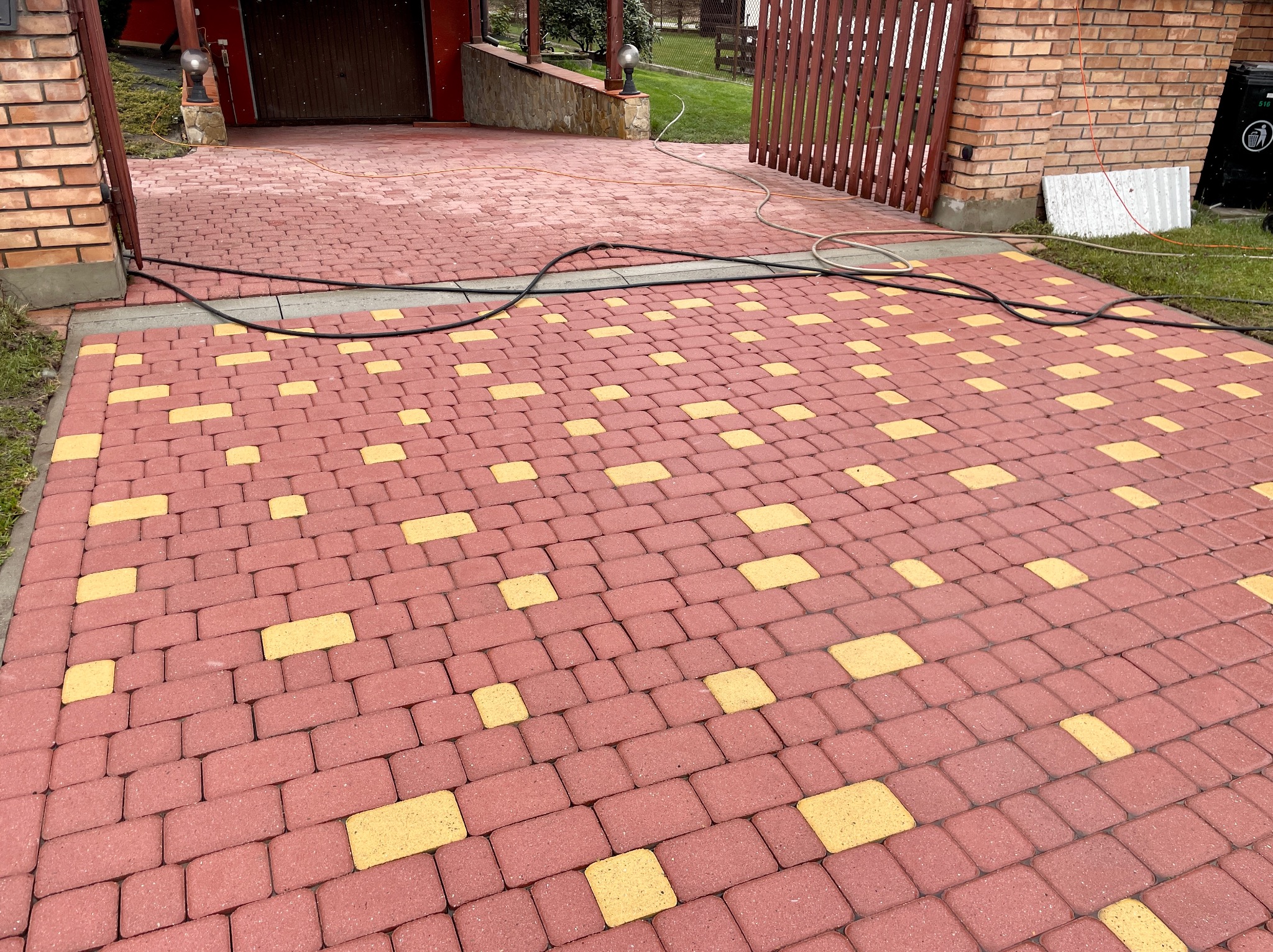 Czerwono-żółta kostka brukowa na podjeździe, widok z góry, z widocznymi przewodami i fragmentem bramy garażowej w tle.