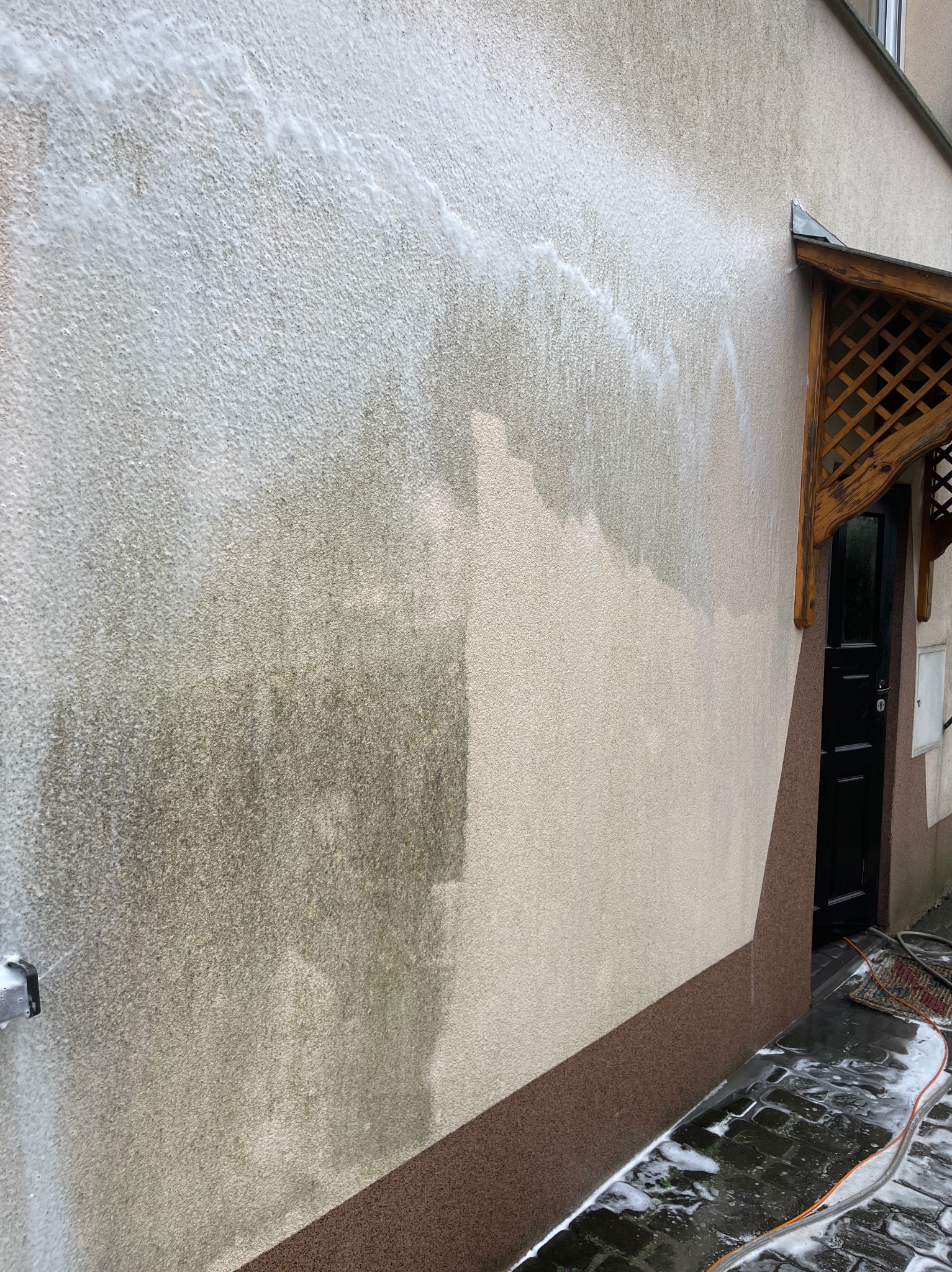 Fragment elewacji budynku w trakcie czyszczenia pod ciśnieniem. Widoczna różnica między umytą a zabrudzoną powierzchnią, piana na kostce brukowej.