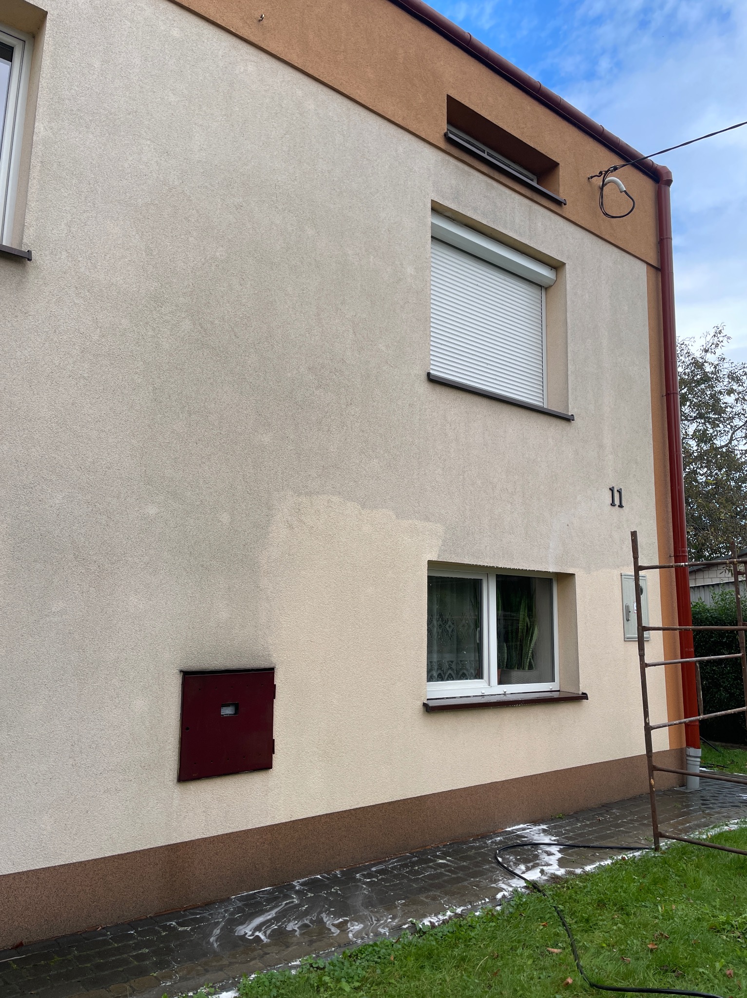 Fragment elewacji budynku z numerem 11, częściowo umyta, ukazująca różnicę w kolorze. Widoczne okna, roleta, skrzynka i fragment kostki brukowej z resztkami piany.