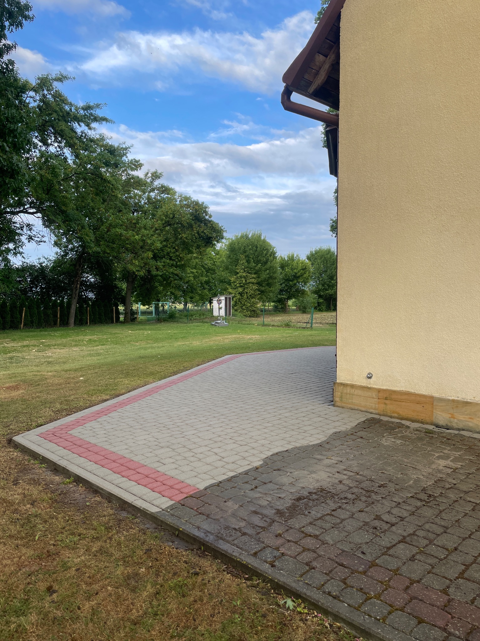 Jasnoszara kostka brukowa z czerwoną obwódką, częściowo mokra, przylegająca do beżowej elewacji budynku. W tle trawnik i drzewa pod błękitnym niebem.
