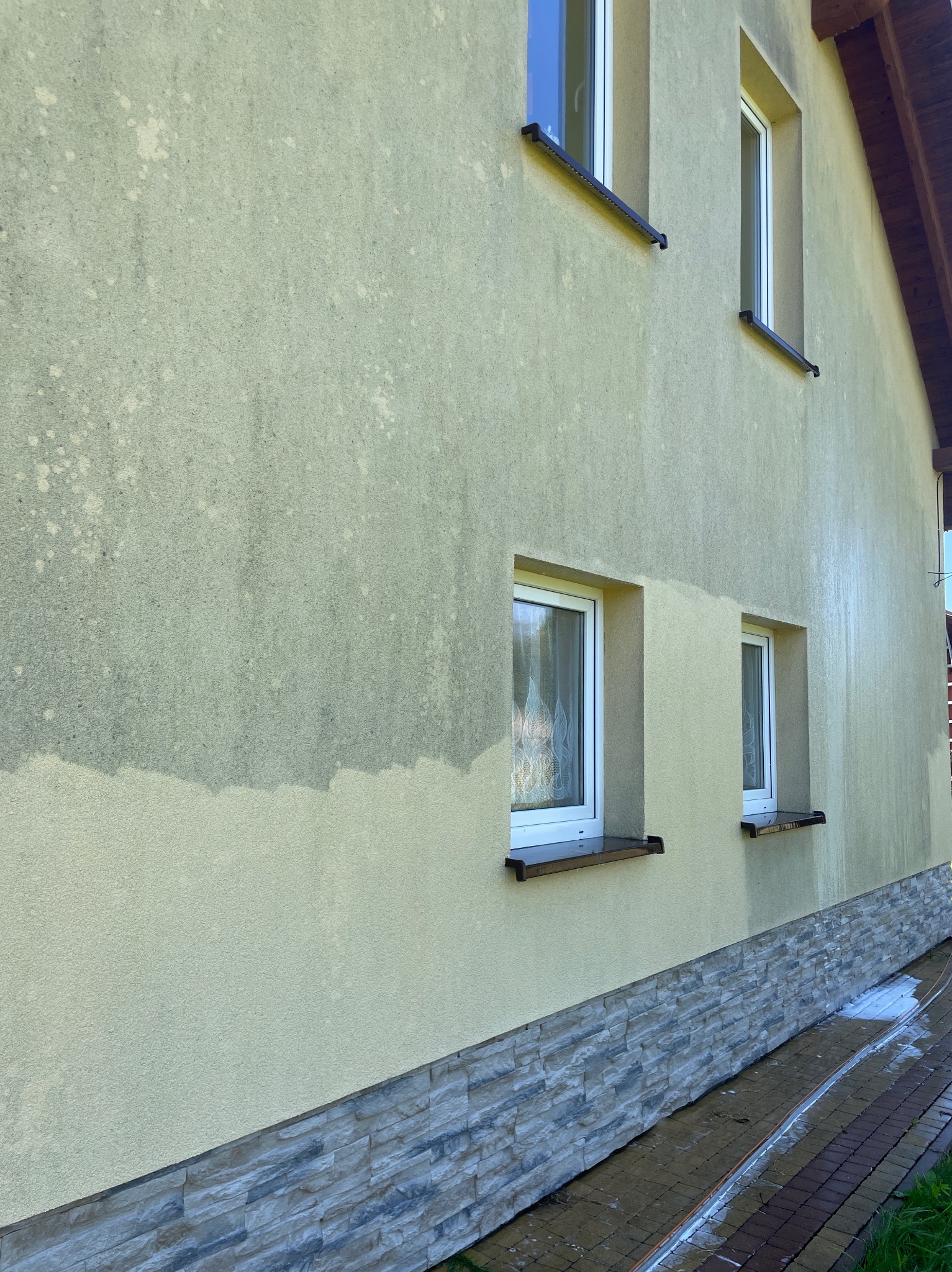 Fragment elewacji domu z widoczną różnicą po czyszczeniu, od dołu do połowy ściany. Widoczne okna i kamienna podmurówka. Ślady wody na kostce brukowej.