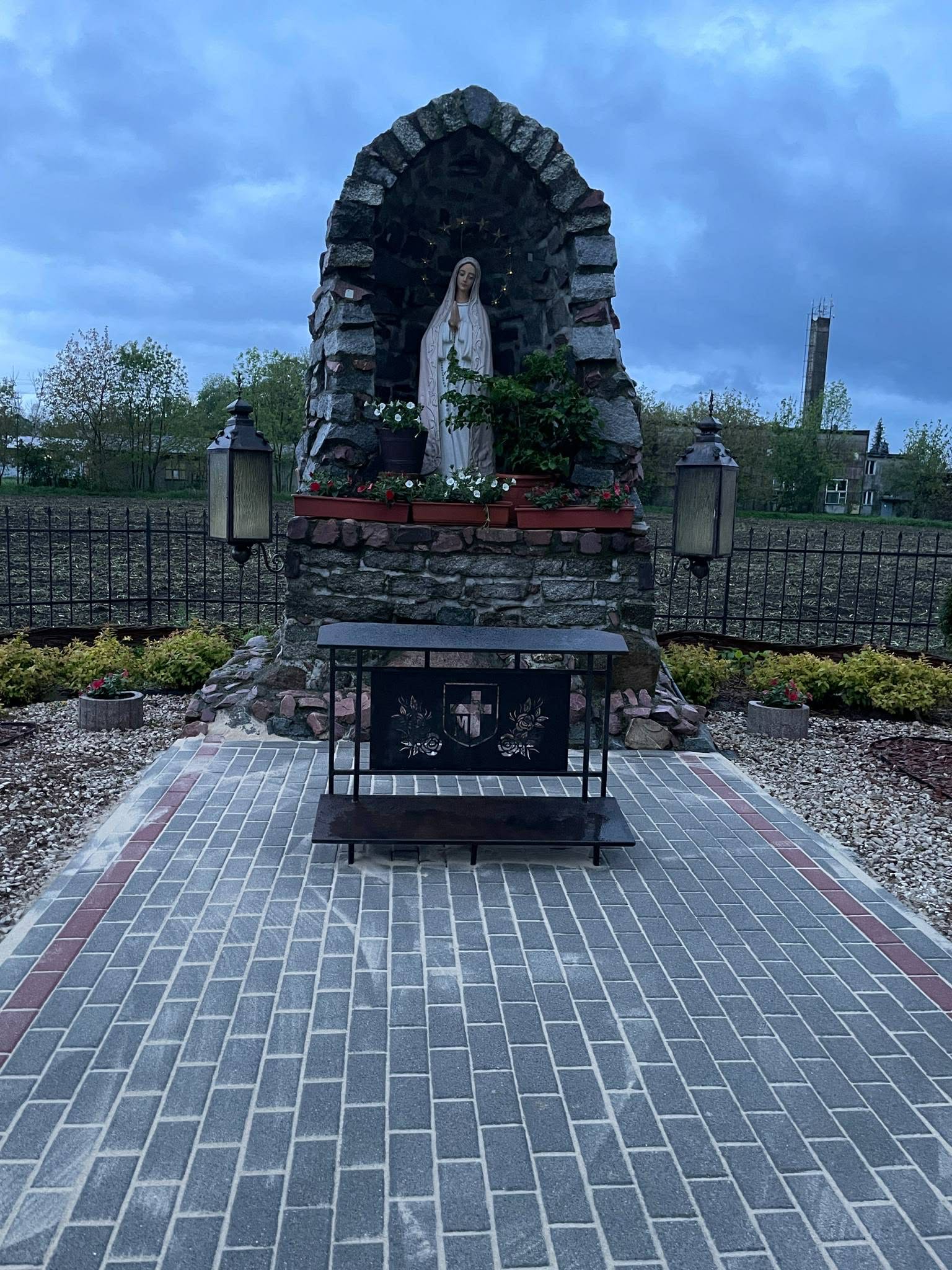 Kapliczka z figurą Matki Boskiej, otoczona kostką brukową i zielenią, oświetlona latarniami. Widok na zadbane otoczenie i pochmurne niebo.