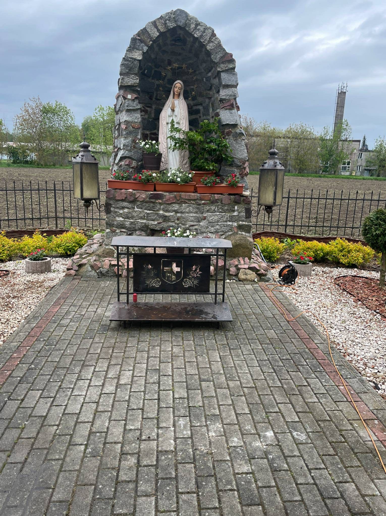 Kapliczka z figurą Matki Boskiej, otoczona kostką brukową i zielenią. Widoczne lampy, kwiaty w donicach i elementy dekoracyjne. W tle pole i komin.
