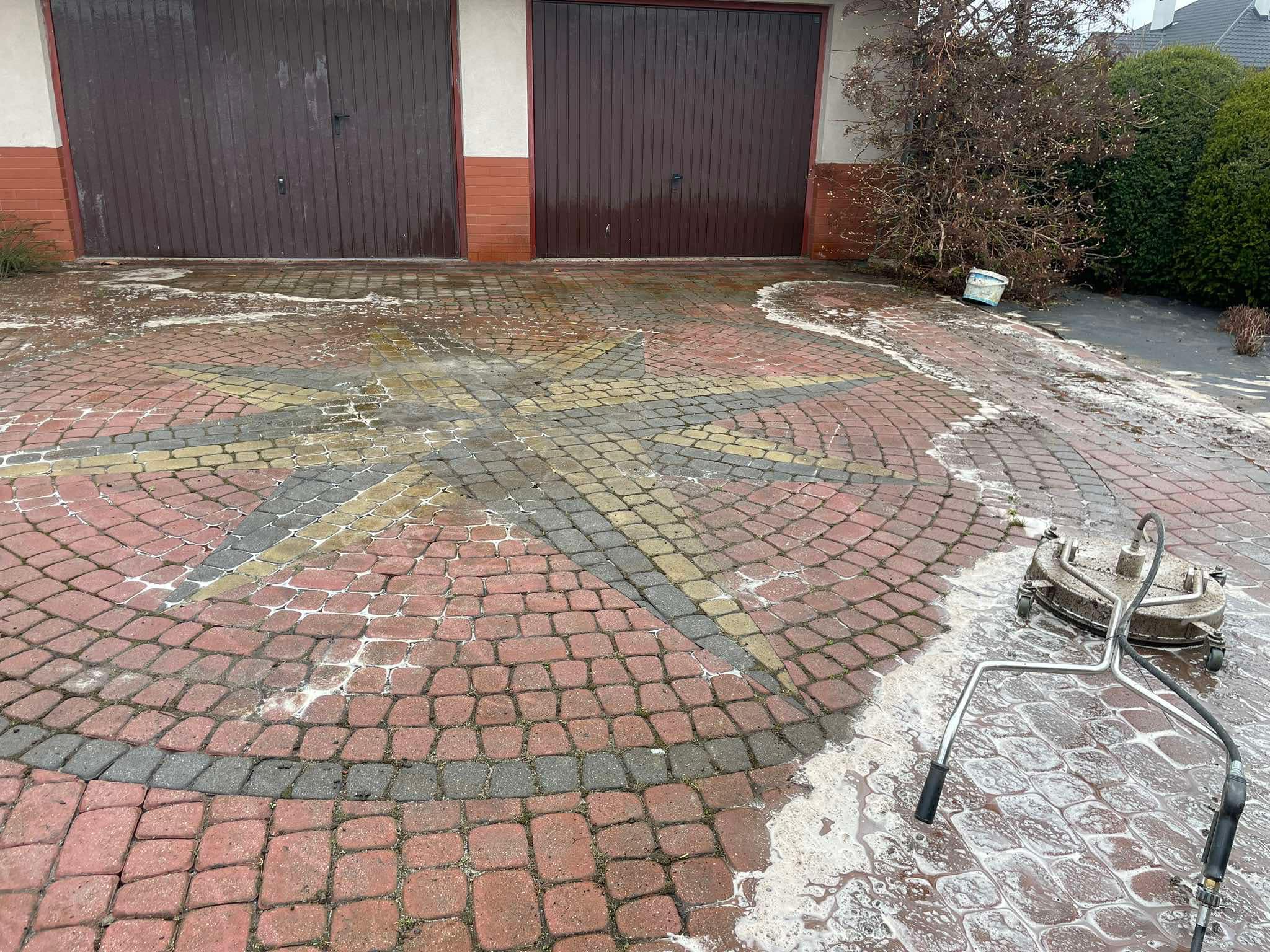Mycie kostki brukowej z wzorem gwiazdy w Przemyślu. Częściowo umyta powierzchnia z użyciem myjki ciśnieniowej. Widoczne dwie bramy garażowe w tle.