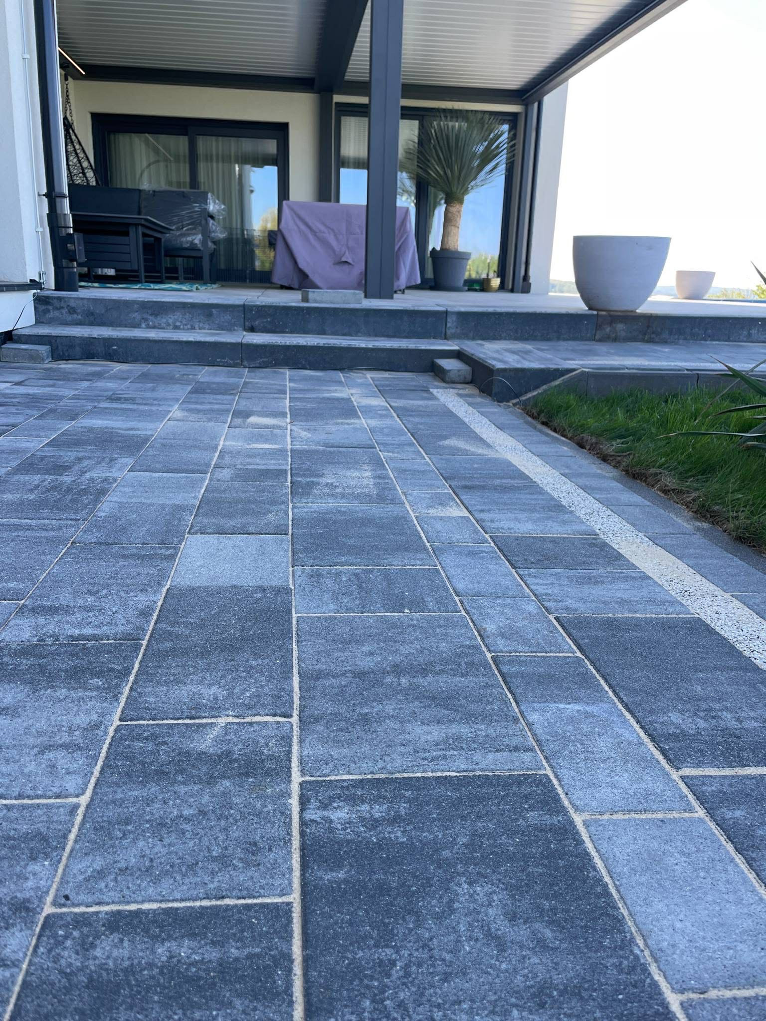 Ułożona, szara kostka brukowa na posesji z elementami zieleni i tarasu. Widok z niskiej perspektywy podkreśla regularność wzoru i estetykę wykończenia.