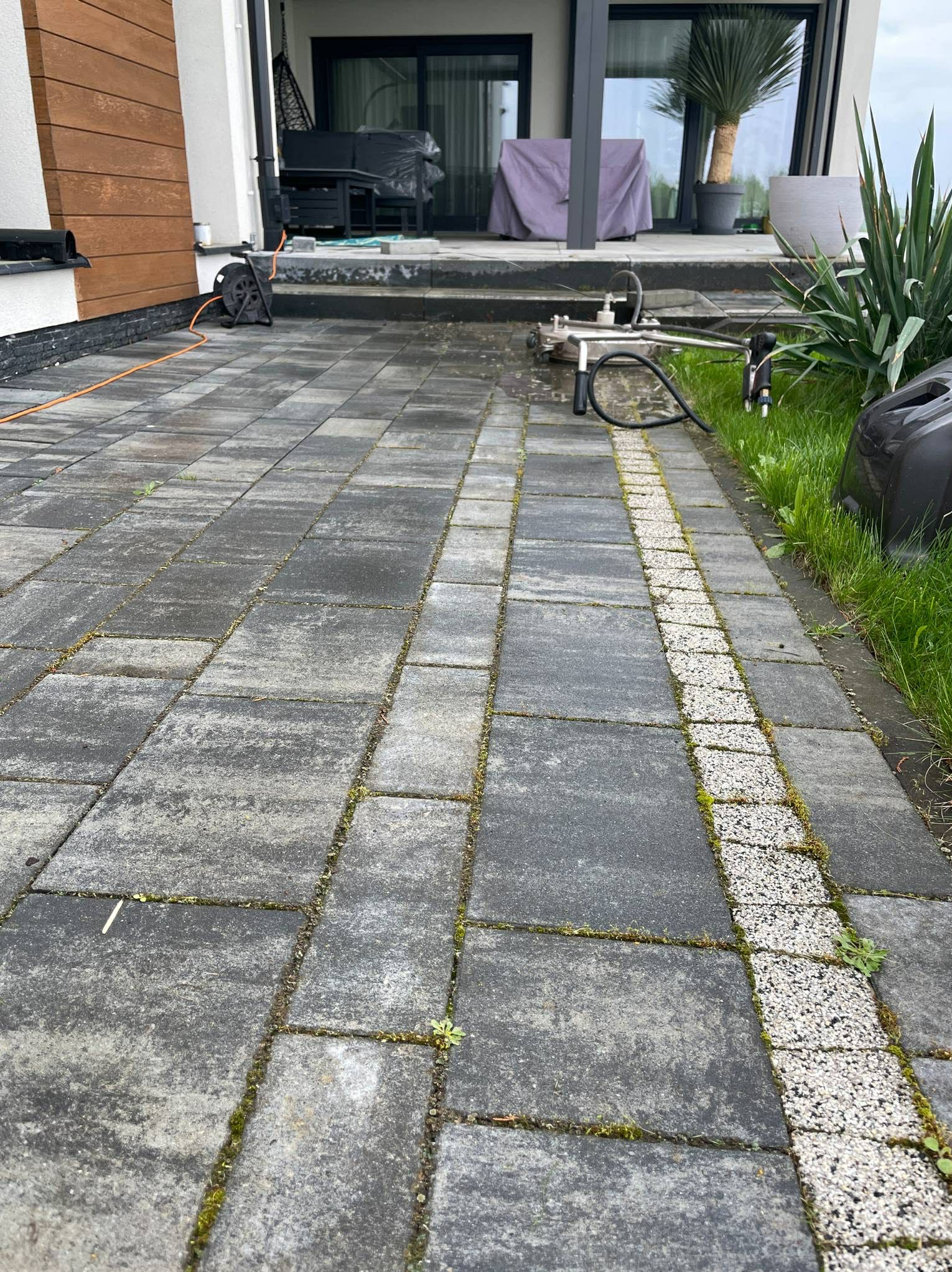 Szara kostka brukowa z mchem w szczelinach, częściowo umyta. W tle narzędzie do mycia pod ciśnieniem, zwinięty kabel i rośliny w donicach przy nowoczesnym domu.