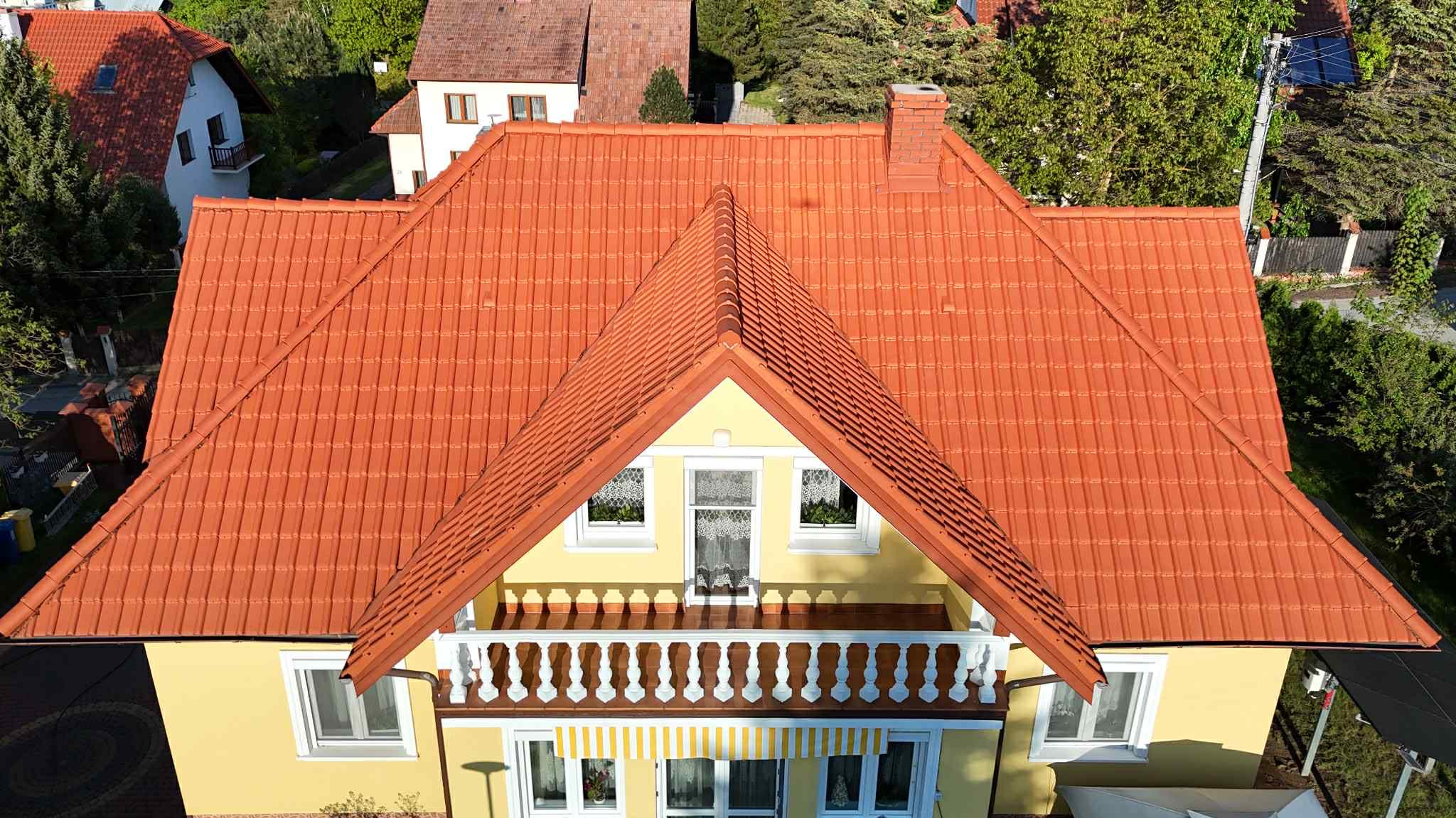 Żółty dom z nową, czerwoną dachówką, widok z góry. Dachówka ceramiczna, symetryczny układ, białe okna i balkon. Dom otoczony zielenią.