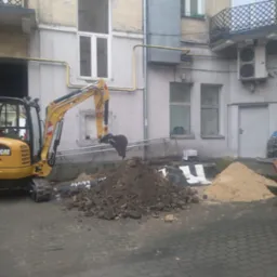 Żółta minikoparka CAT 302.7D nabiera ziemię na tle budynku z oknami i balkonem, obok leżą pryzmy ziemi i piasku.